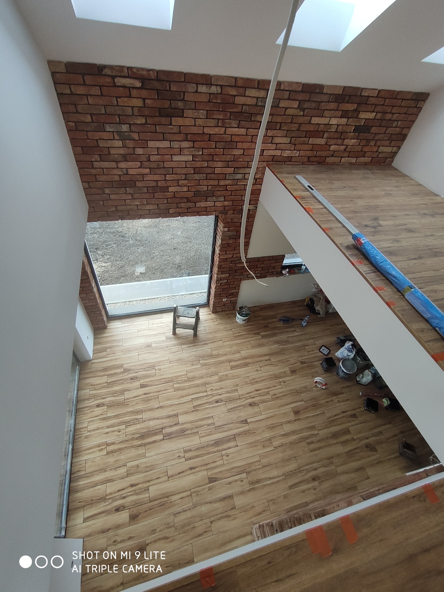 Wnętrze domu w trakcie prac wykończeniowych: ściana z cegły klinkierowej, drewnopodobna podłoga, okno dachowe, widoczne narzędzia i puszki po farbach, niebieska folia ochronna na schodach.