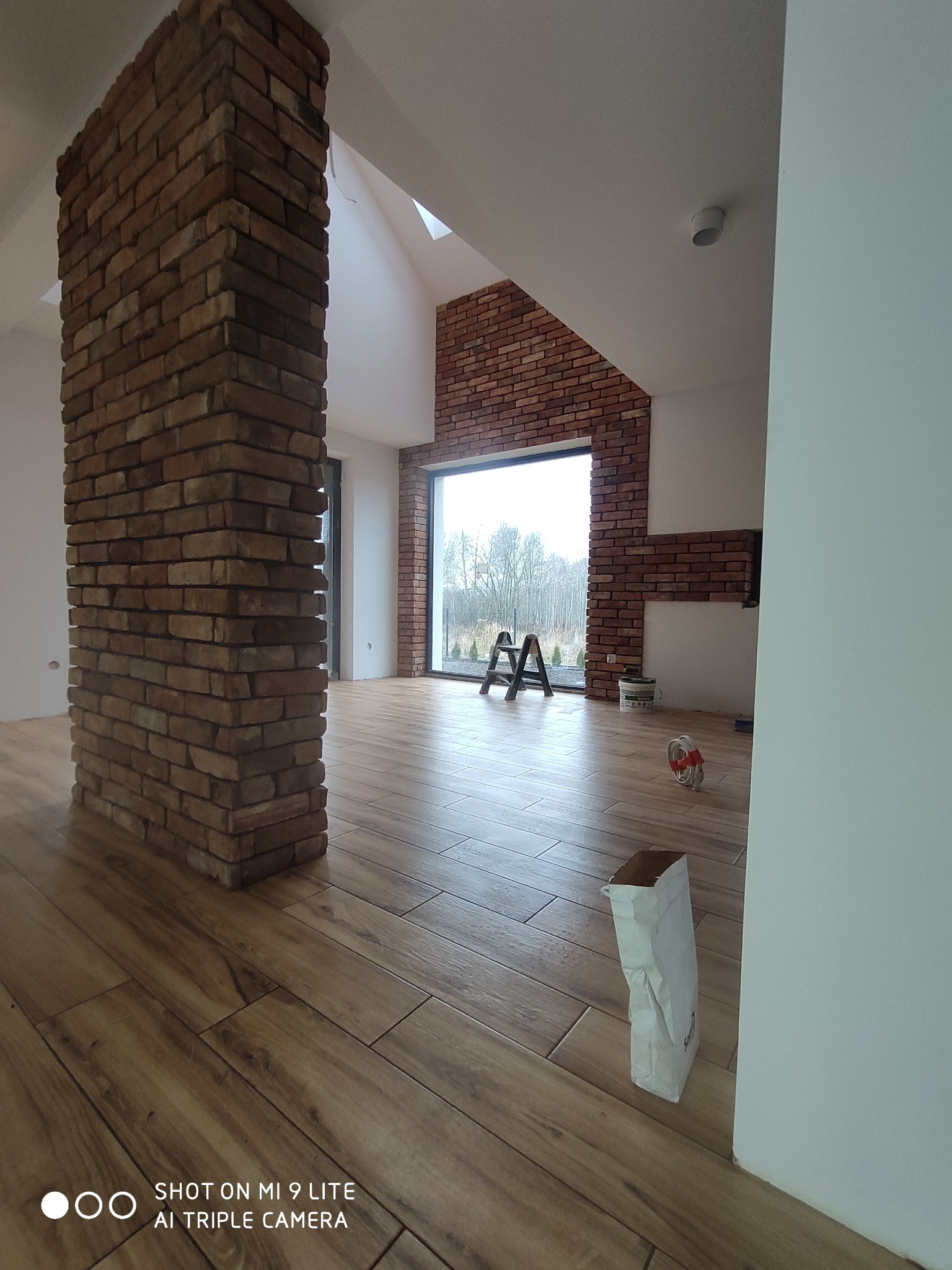 Wnętrze w trakcie wykańczania z kolumną obłożoną cegłą klinkierową, drewnopodobną podłogą, ścianą z cegły przy oknie i workiem zaprawy na podłodze.