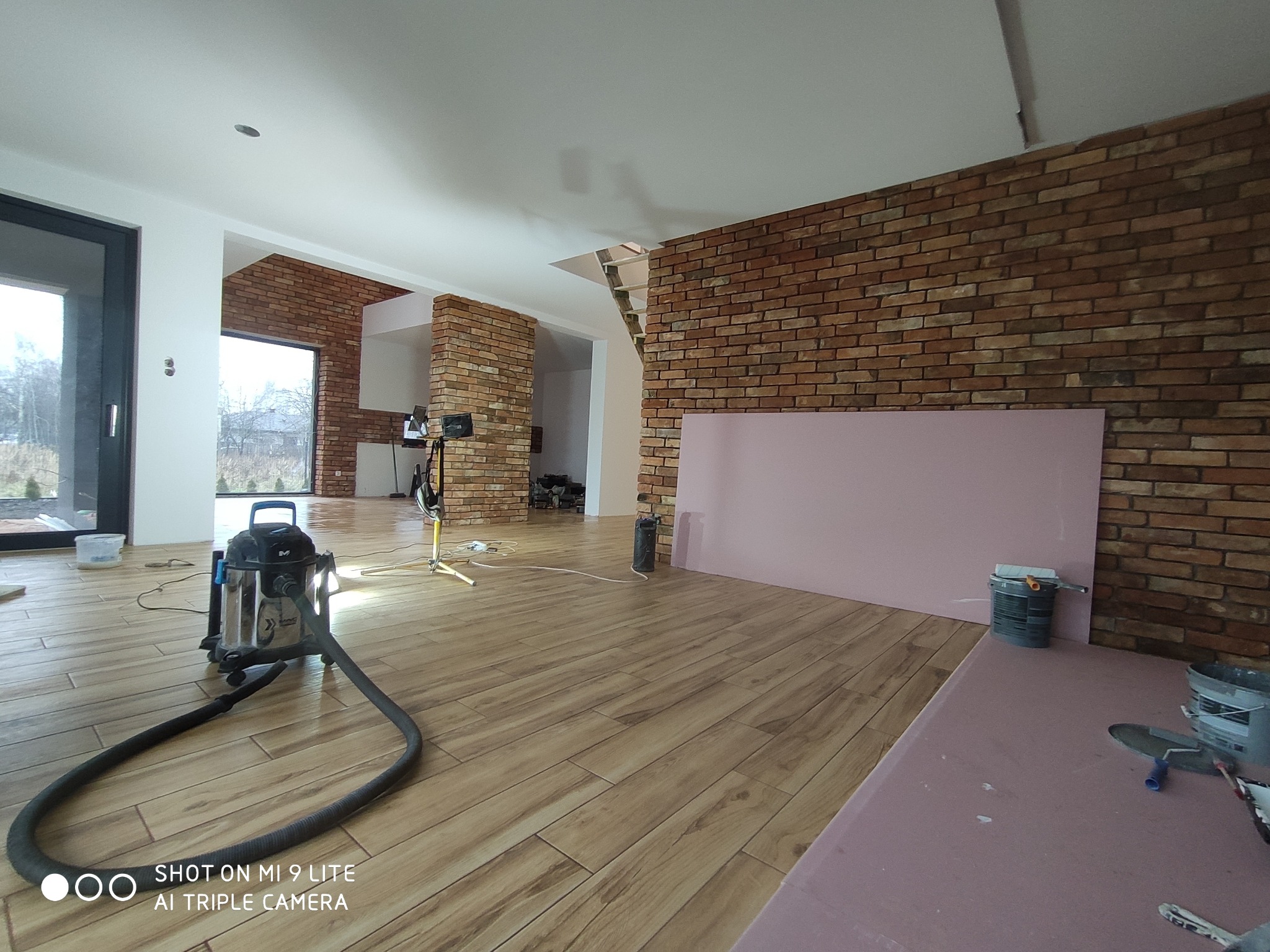 Wnętrze w trakcie prac wykończeniowych: widoczny odkurzacz przemysłowy, fragment ściany z cegły, płyty kartonowo-gipsowe w kolorze różowym, podłoga wyłożona panelami imitującymi drewno,...