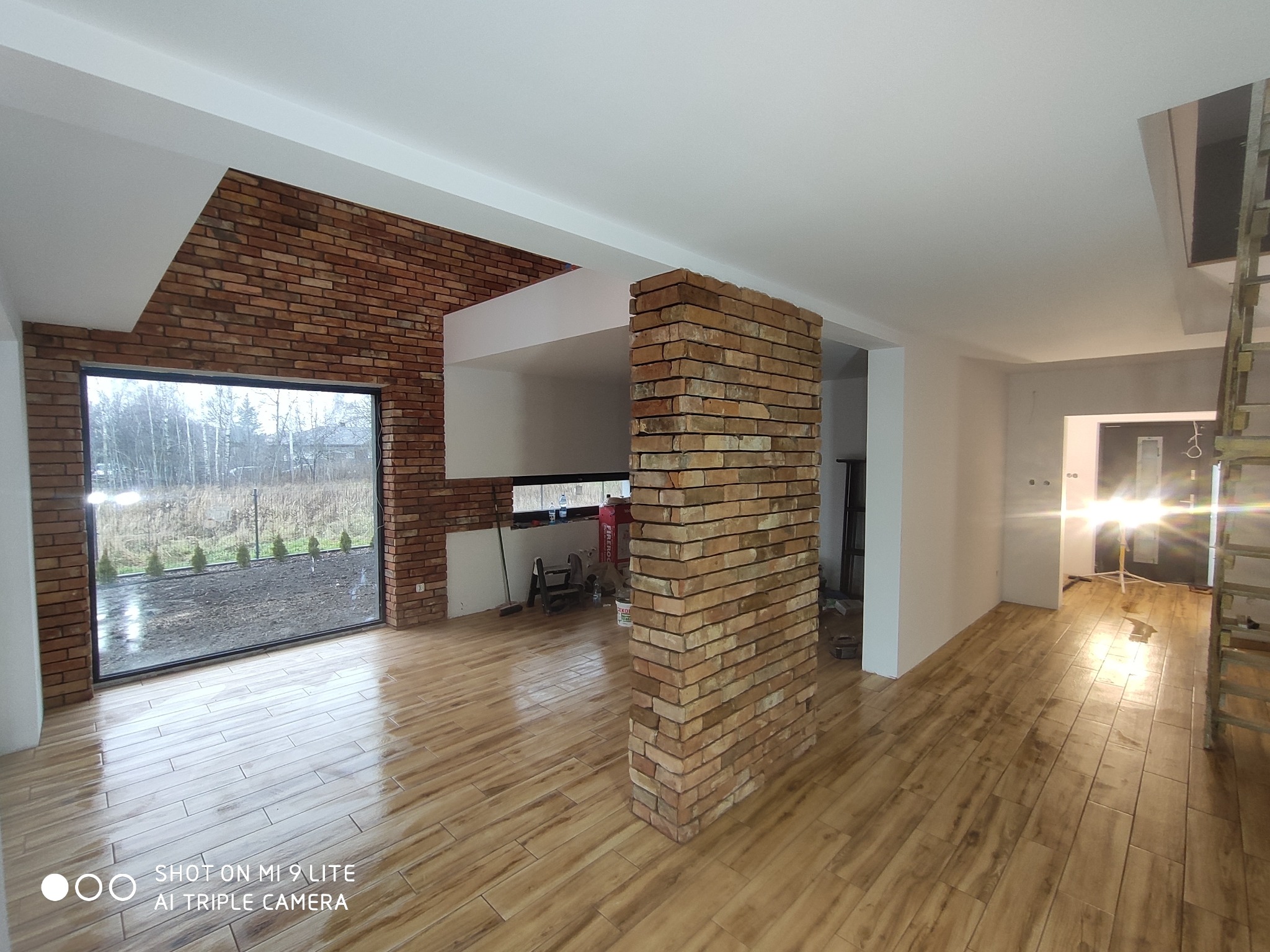 Wnętrze domu w trakcie wykańczania, z ceglaną ścianą i filarem, oraz podłogą imitującą drewno, widok na zewnątrz przez duże okno.