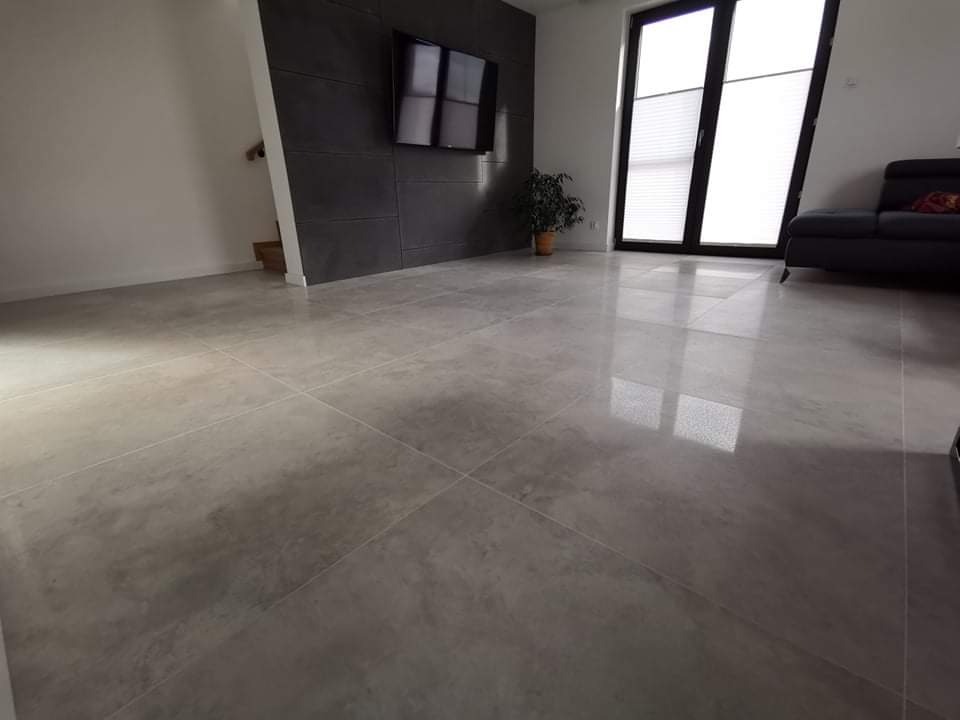 Wyłożona jasnoszarą, błyszczącą płytką podłoga w salonie z ciemną ścianą TV, dużym oknem z żaluzjami i czarną sofą w tle.