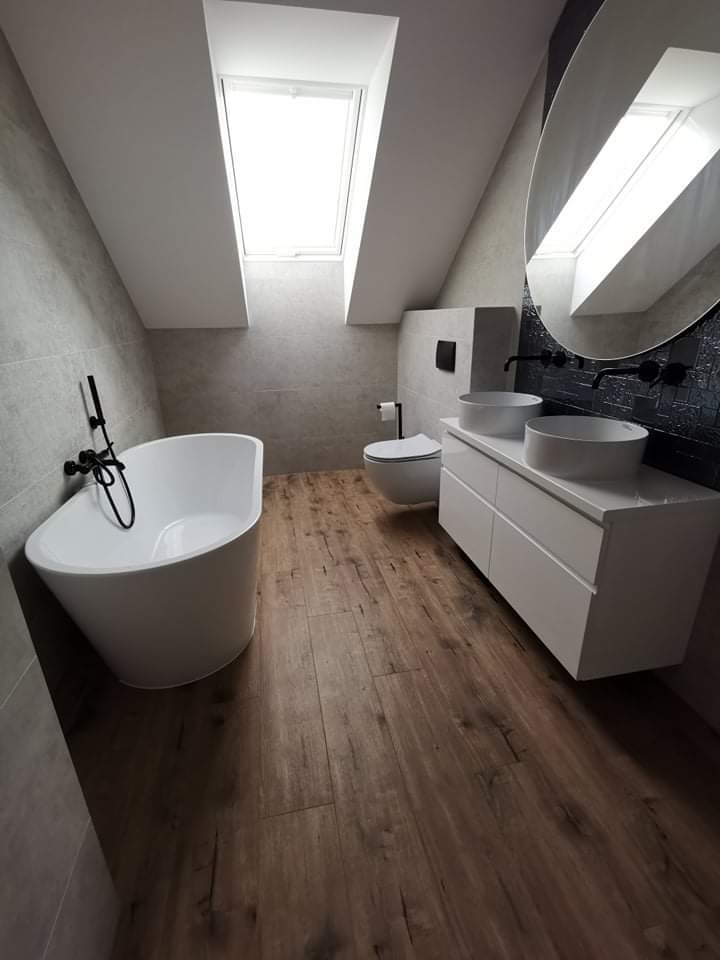 Nowoczesna łazienka na poddaszu z drewnopodobną podłogą, owalną wanną, umywalkami nablatowymi i dużym, okrągłym lustrem.