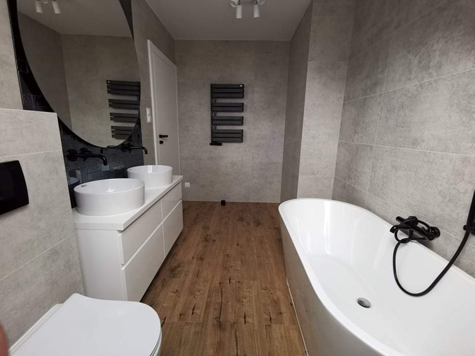 Nowoczesna łazienka w szarościach z drewnopodobną podłogą, owalnym lustrem i wolnostojącą wanną z czarną armaturą.