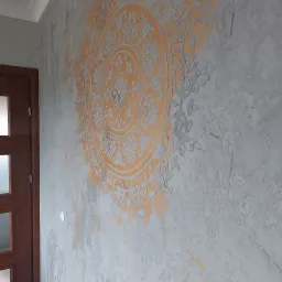 Dekoracyjna ściana z szarą, teksturowaną gładzią i pomarańczowym, okrągłym wzorem przypominającym mandalę, widoczny fragment drewnianej ościeżnicy drzwiowej po lewej stronie.