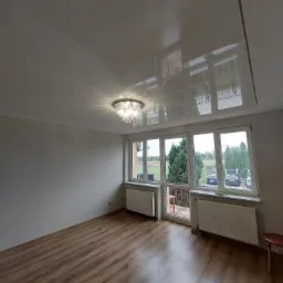 Biały, błyszczący sufit z widocznymi refleksami światła i odbiciami okien w nowo wykończonym pokoju z drewnianą podłogą i ozdobnym żyrandolem.