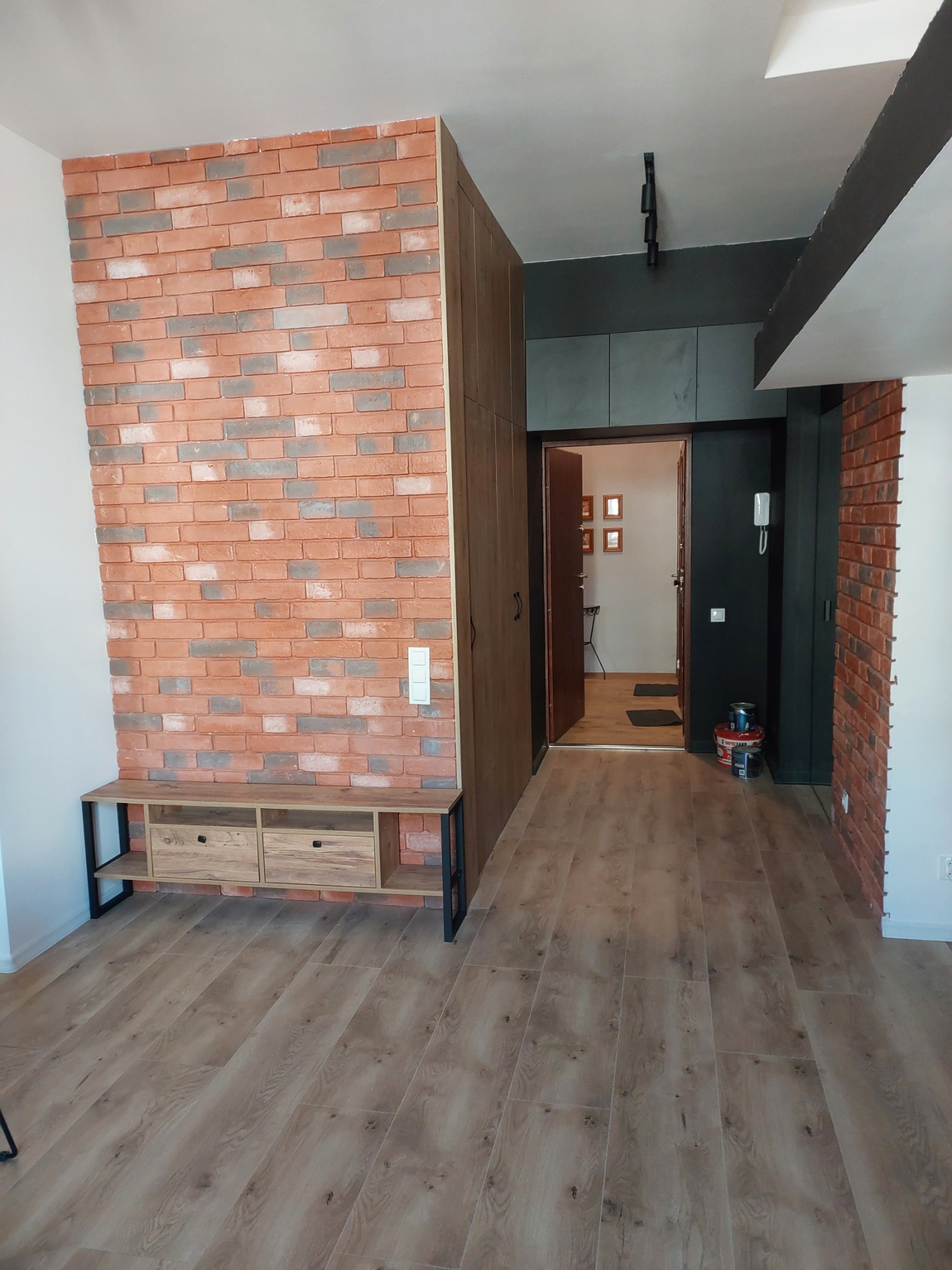 Wnętrze mieszkania z dekoracyjną ścianą z cegły, drewnianą podłogą i meblami w stylu industrialnym, widok na otwarte drzwi do innego pomieszczenia.