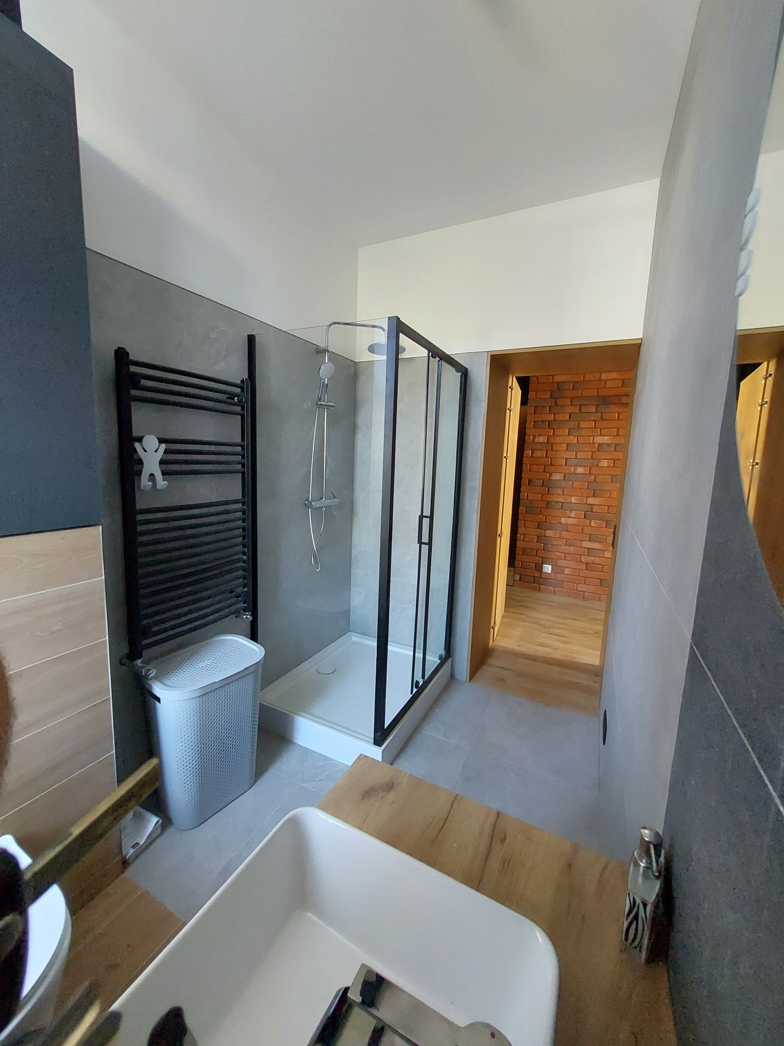Nowoczesna łazienka z kabiną prysznicową typu walk-in, grzejnikiem drabinkowym z wieszakiem w kształcie ludzika, umywalką nablatową i ścianą z cegły w tle.