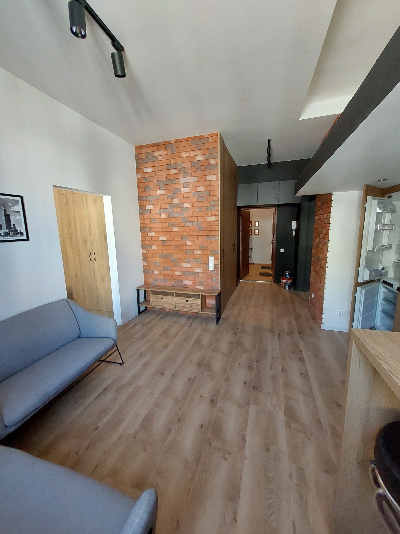 Jasne, przestronne wnętrze mieszkania z podłogą z paneli, ścianą z dekoracyjnej cegły, szarą sofą i oświetleniem szynowym na suficie.