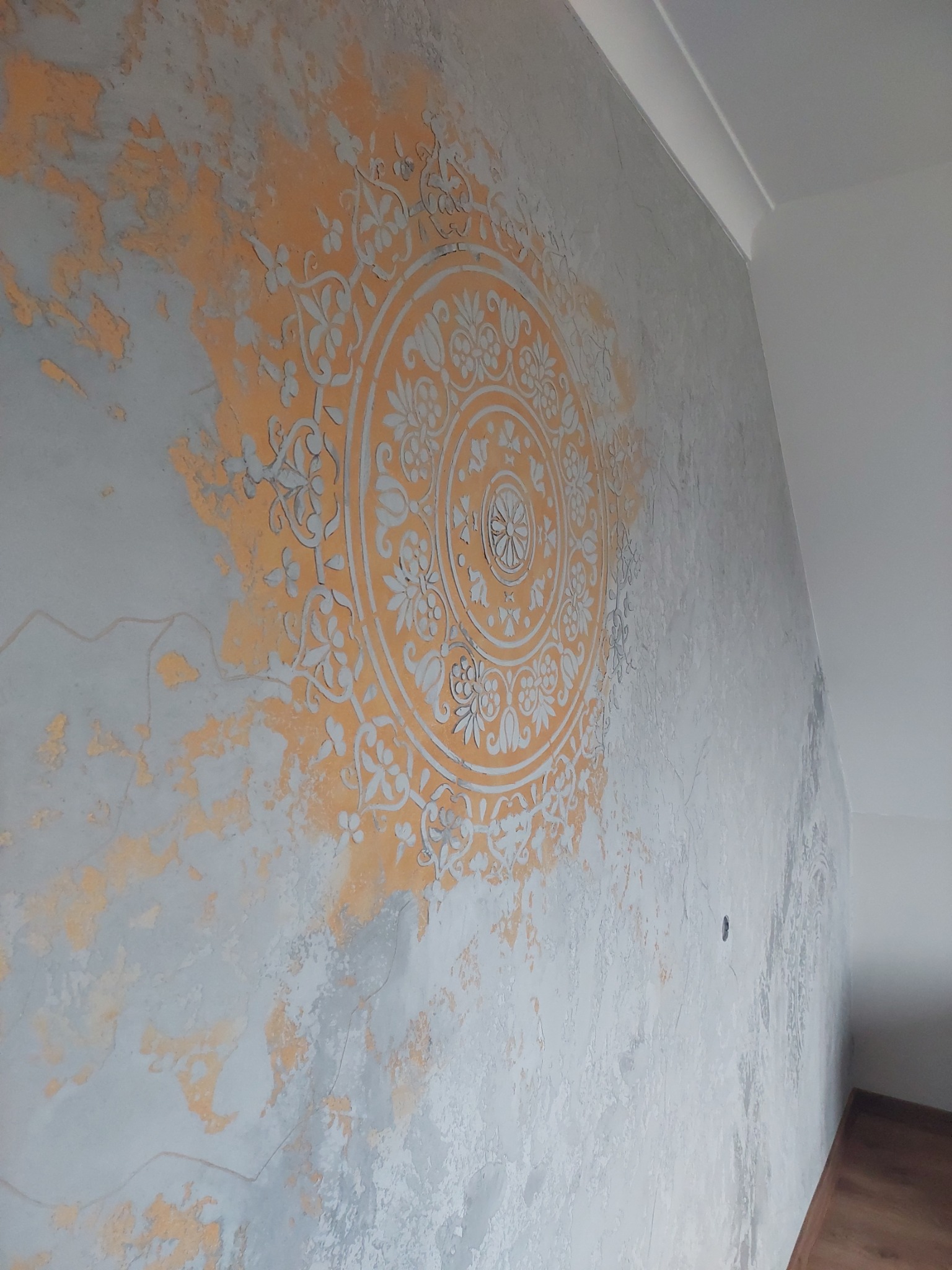 Dekoracyjna ściana z efektem przetarć i ornamentem mandali w odcieniach szarości i pomarańczu, widok z boku.