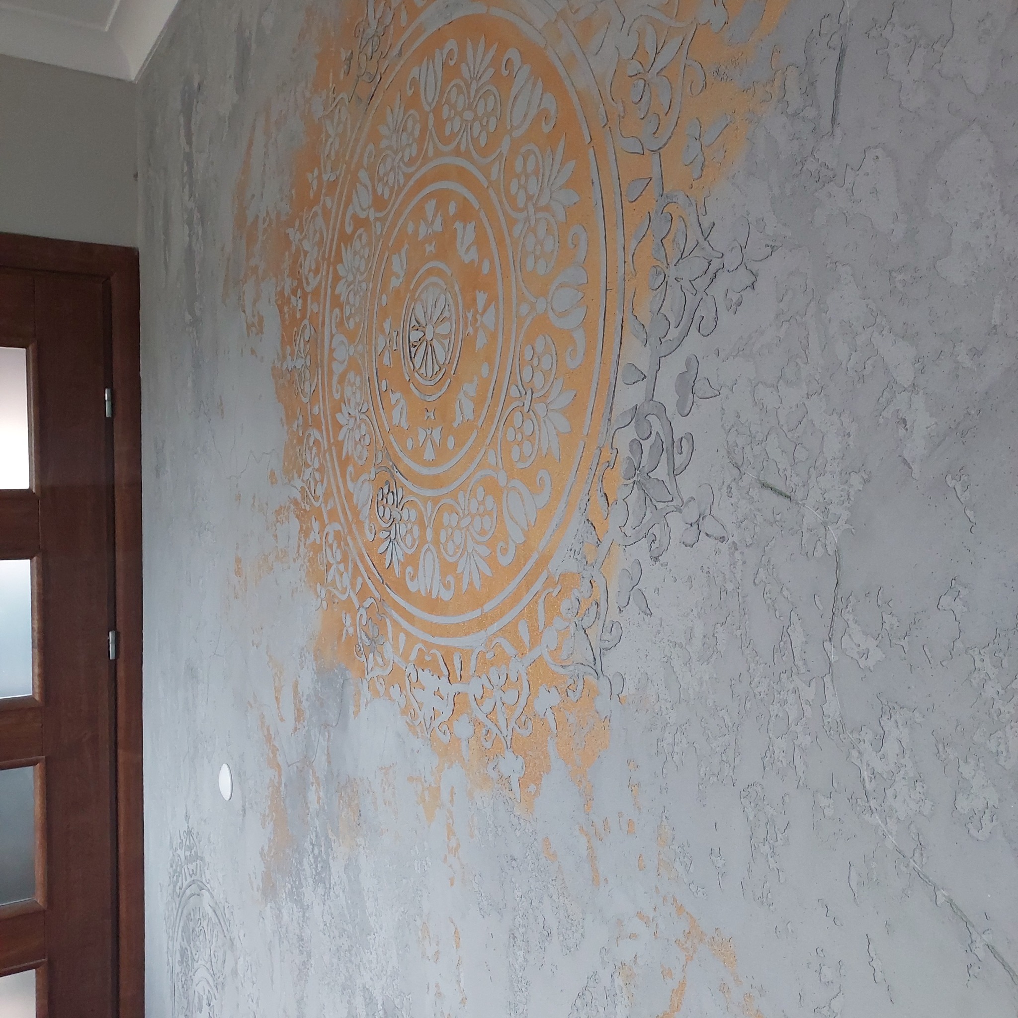 Dekoracyjna ściana z szarą, teksturowaną gładzią i pomarańczowym, okrągłym wzorem przypominającym mandalę, widoczny fragment drewnianej ościeżnicy drzwiowej po lewej stronie.