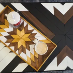 Drewniany stół z geometrycznym wzorem chevron w odcieniach bieli, brązu i czerni, na którym stoi taca z identycznym wzorem, a na niej filiżanka kawy i dzbanek do herbaty.