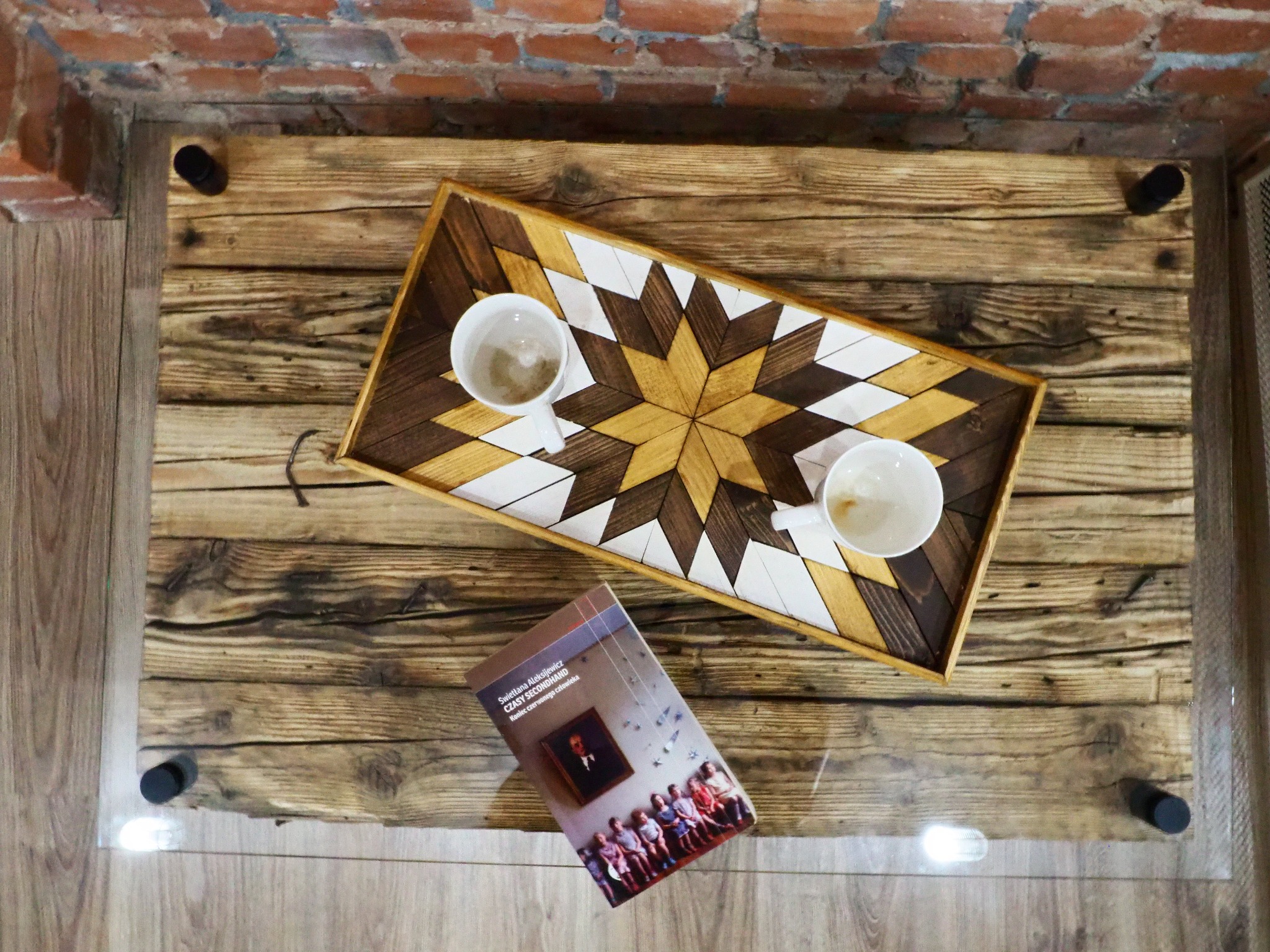 Widok z góry na drewniany stolik kawowy z blatem z desek i szklaną powierzchnią, na którym leży taca z geometrycznym wzorem gwiazdy, dwie filiżanki po kawie i książka Swietłany Aleksijewicz 'Czasy...