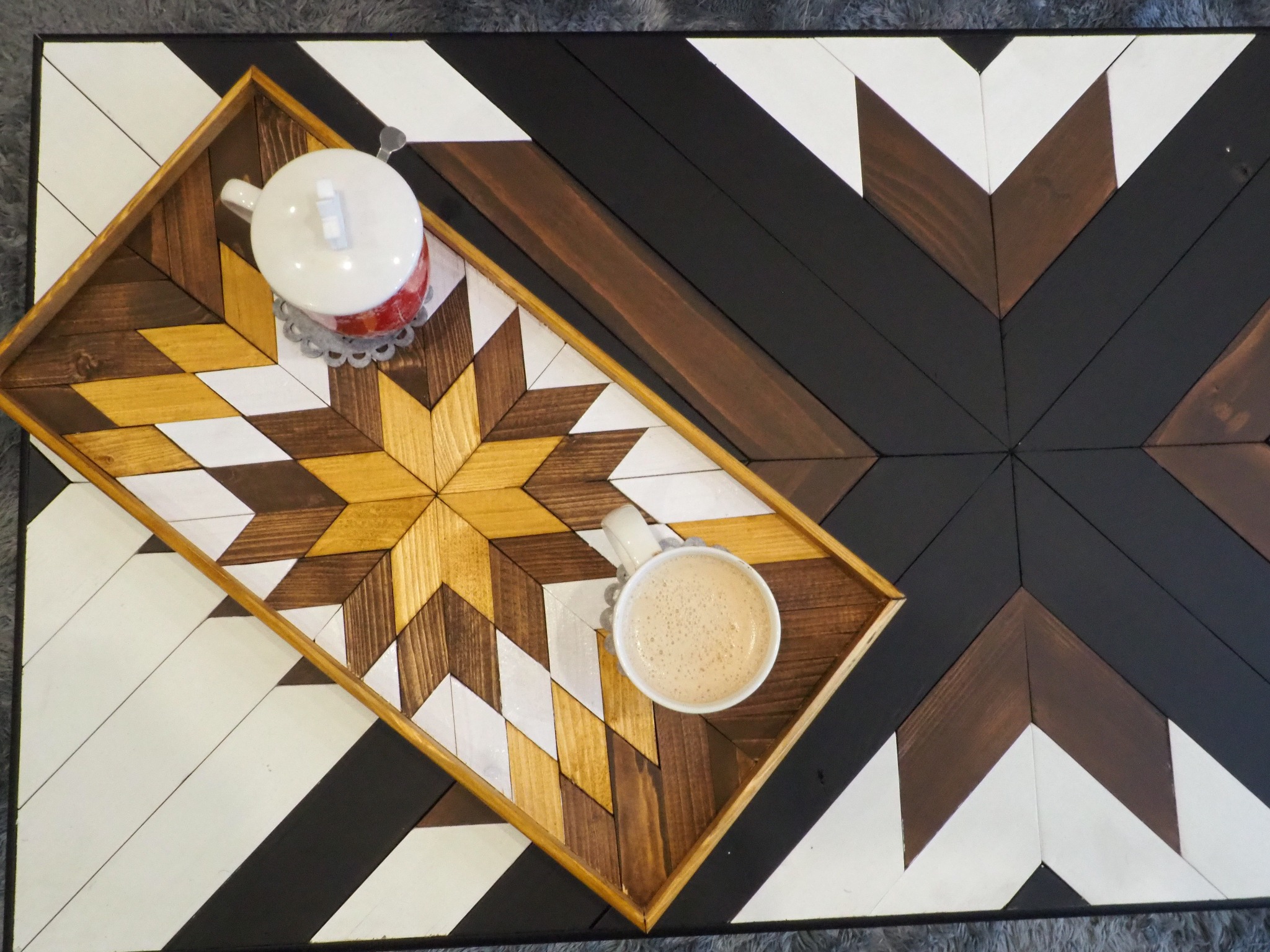 Drewniany stół z geometrycznym wzorem chevron w odcieniach bieli, brązu i czerni, na którym stoi taca z identycznym wzorem, a na niej filiżanka kawy i dzbanek do herbaty.