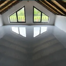 Świeżo wylana, szara, błyszcząca podłoga w pomieszczeniu na poddaszu z dwoma tr&oacute;jkątnymi oknami, odbijającymi się w powierzchni podłogi.