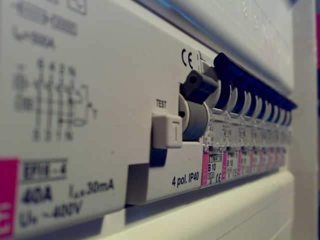 Rozdzielnica elektryczna z widocznymi bezpiecznikami, etykietami i schematem, oświetlona z boku, ukazująca stan załączenia jednego z wyłączników.