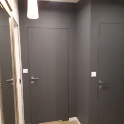 Nowoczesne, szare drzwi ukryte w ścianie, z minimalistycznymi klamkami i włącznikami, drewniana podłoga w trakcie zabezpieczania folią malarską.