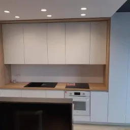 Nowoczesna kuchnia z białymi, matowymi frontami szafek, drewnianym blatem i zabudową piekarnika, uzupełniona oświetleniem punktowym i wysoką szafą.