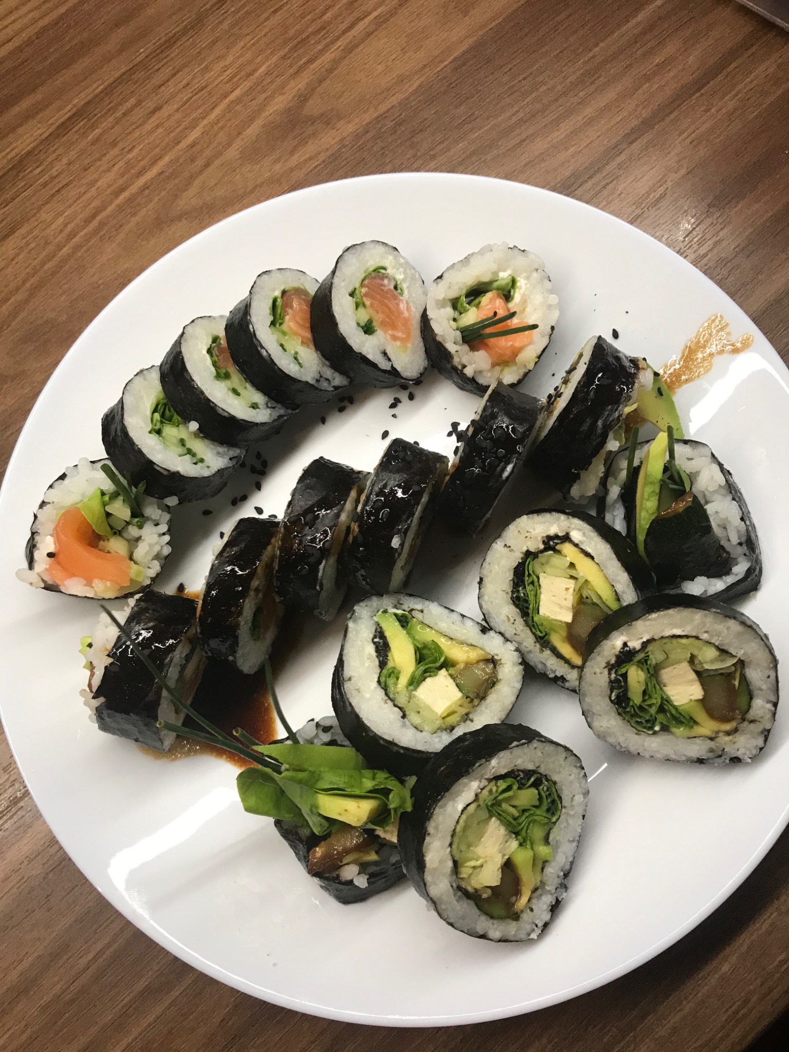 Różnorodny zestaw sushi na białym talerzu, z rolkami z łososiem i wegańskimi z tofu, udekorowany sosem sojowym i świeżymi ziołami, na drewnianym stole.