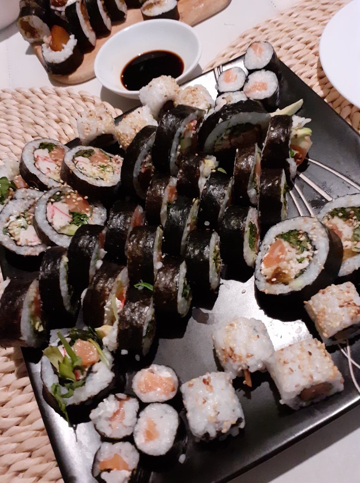 Różnorodny zestaw sushi i maki na czarnym talerzu, z widoczną miseczką sosu sojowego i plecioną podkładką, ukazujący bogactwo smaków i tekstur.