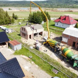 Widok z góry na budowę domu jednorodzinnego z betoniarką podającą beton na dach, w tle krajobraz z jeziorem i lasem.