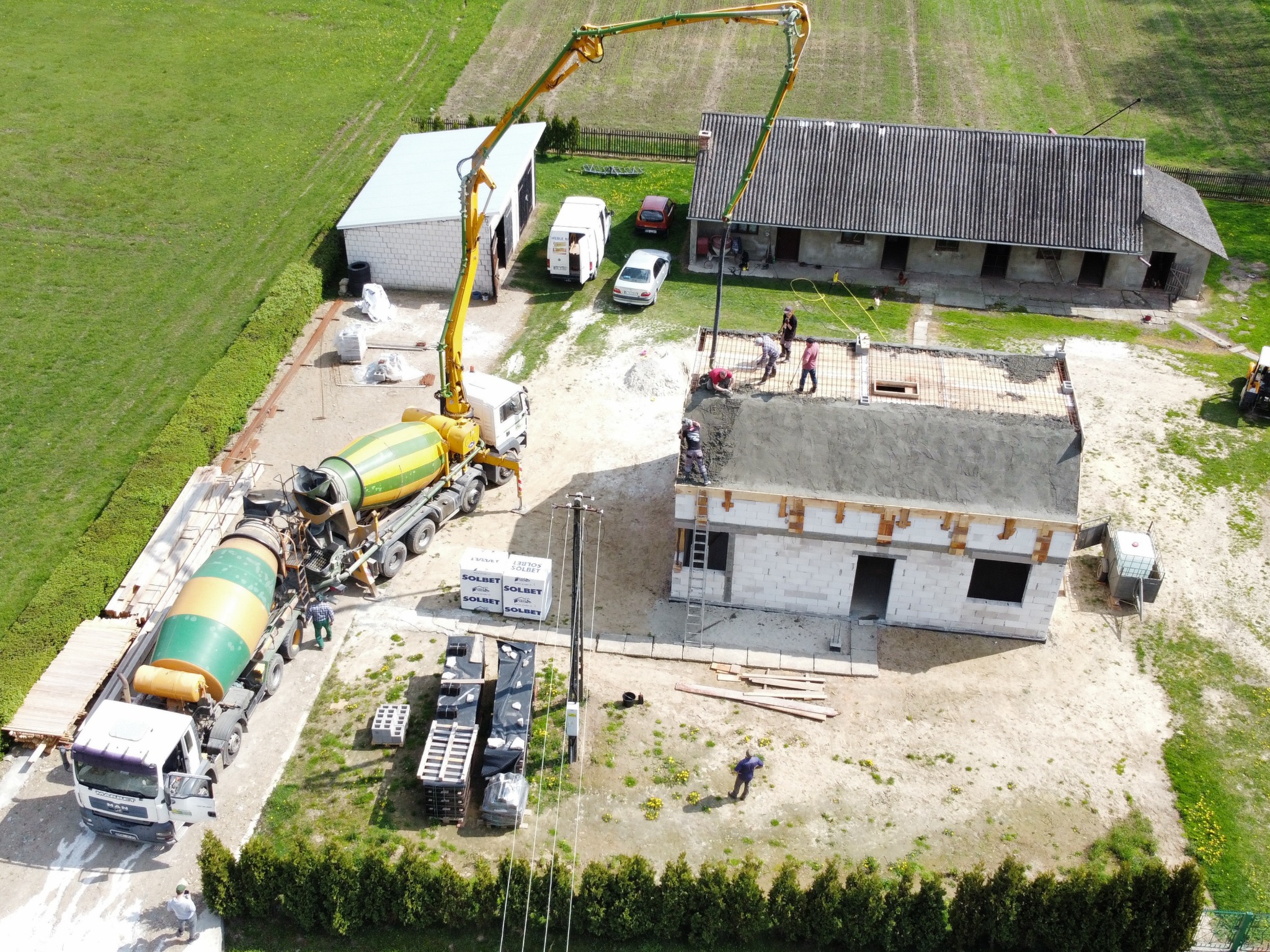 Widok z góry na budowę domu jednorodzinnego, gdzie betonomieszarka z pompą podaje zaprawę na strop. W tle zabudowania gospodarcze i zielone pola.