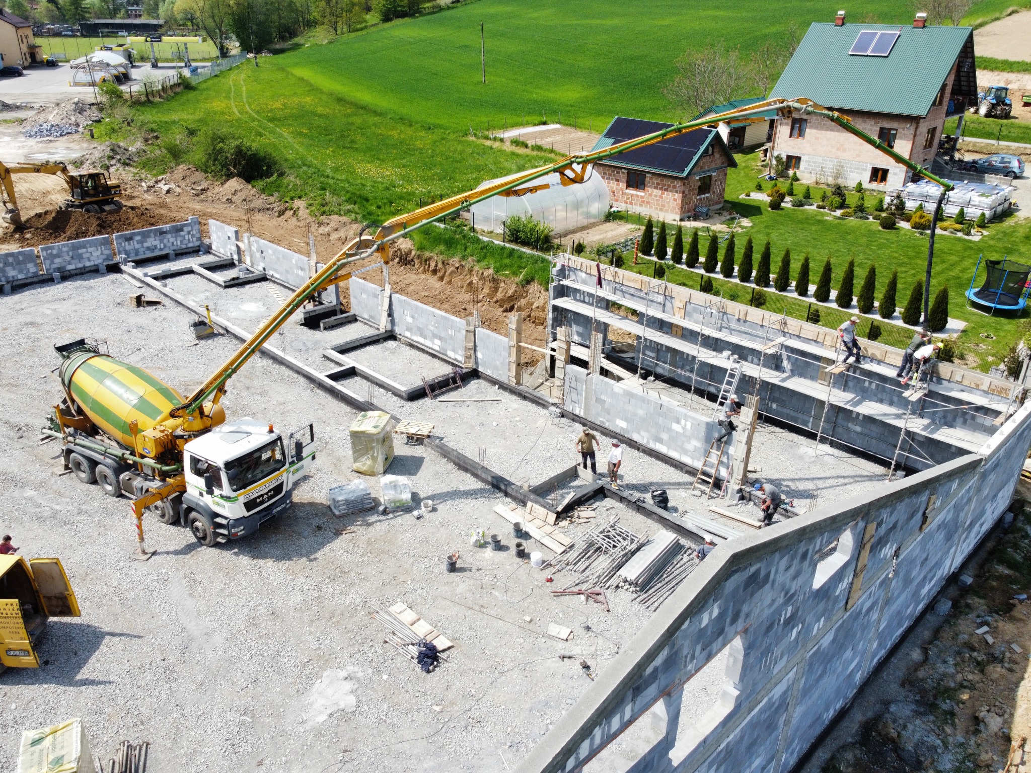 Wysoki kąt widzenia na budowę fundamentów domu jednorodzinnego z szarych bloczków, z użyciem pompy do betonu z ciężarówki w żółto-zielonych barwach. W tle widoczny dom i zielony krajobraz.