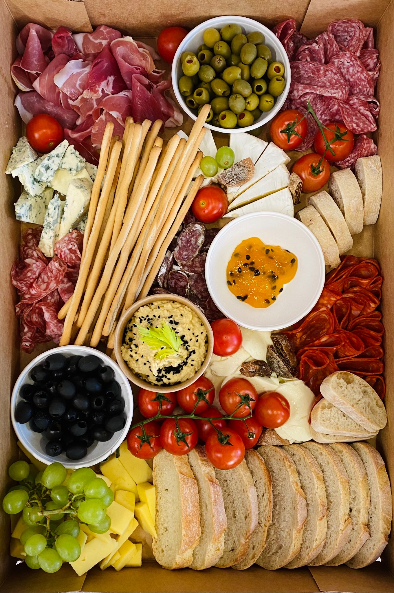 Kompozycja różnorodnych przekąsek w kartonowym pudełku: wędliny, sery pleśniowe i żółte, oliwki zielone i czarne, grissini, hummus z czarnuszką, dżem z marakui, winogrona i pomidorki koktajlowe.