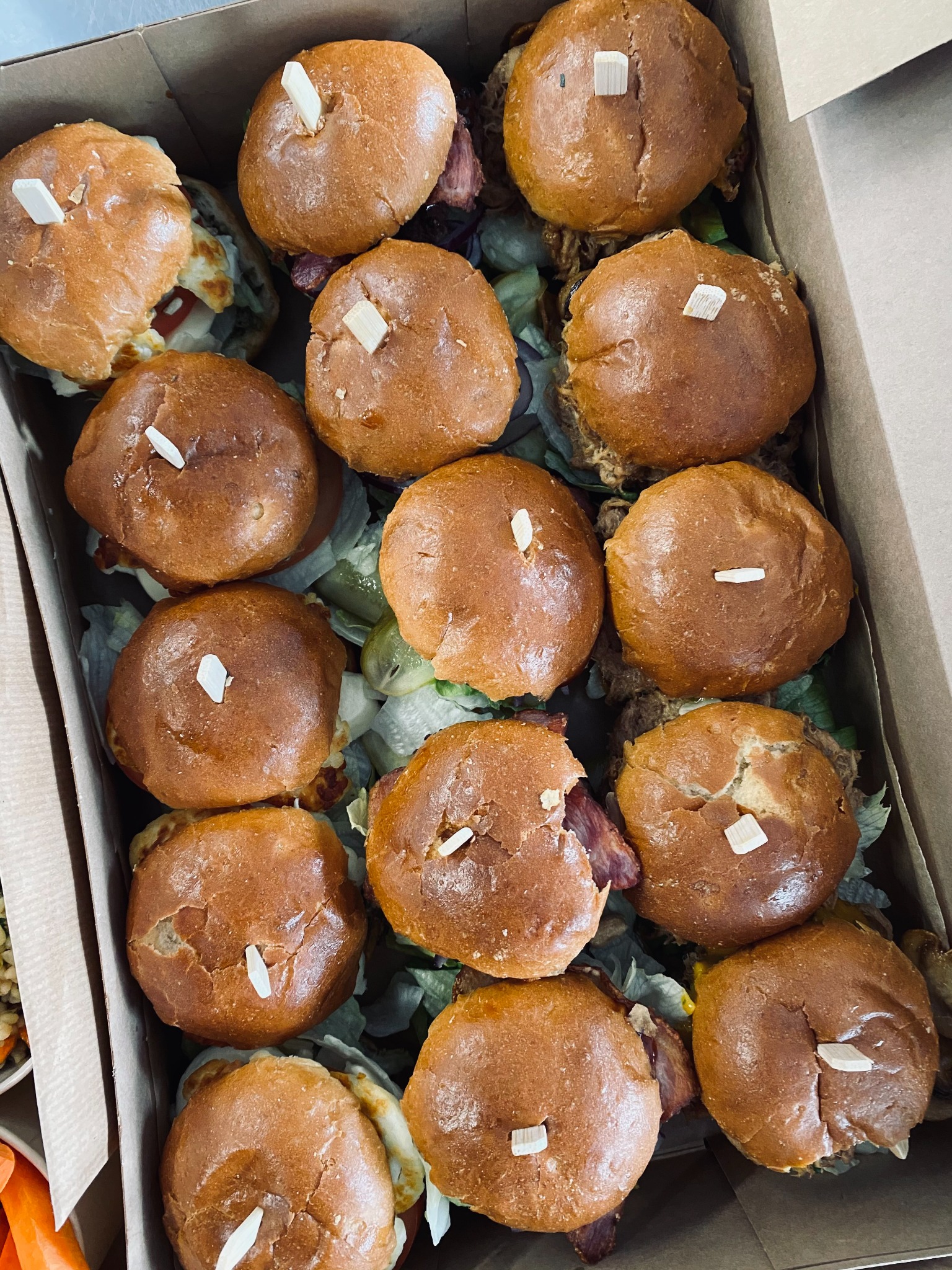 Kartonowe pudełko wypełnione rzędami mini burgerów z drewnianymi wykałaczkami, widoczne składniki: sałata, bekon, ser i sos