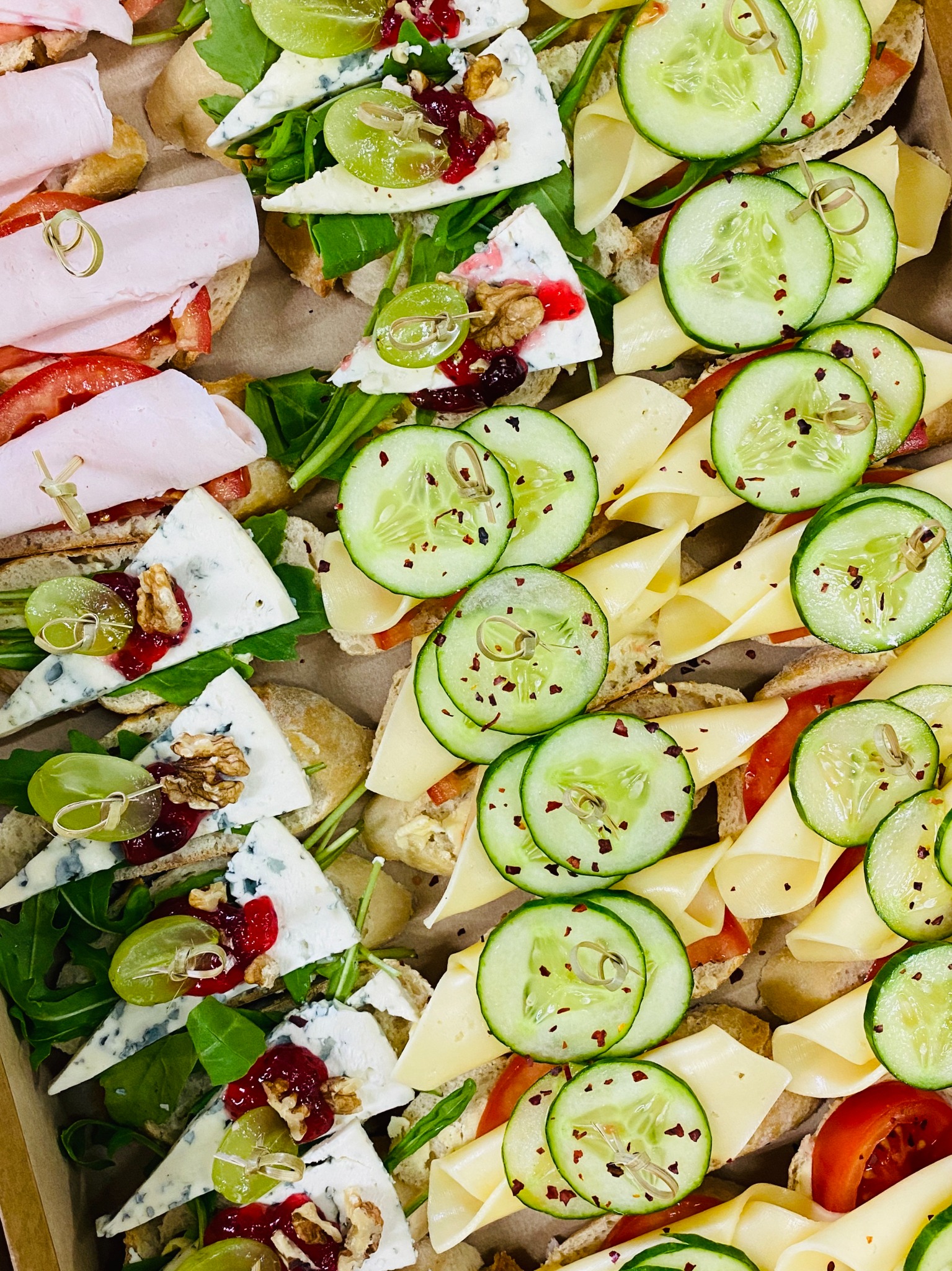 Różnorodne kanapki na jasnym pieczywie: z szynką i pomidorem, serem pleśniowym, winogronem i orzechami włoskimi oraz z żółtym serem i plastrami ogórka posypanymi przyprawami, ułożone w kartonowym...