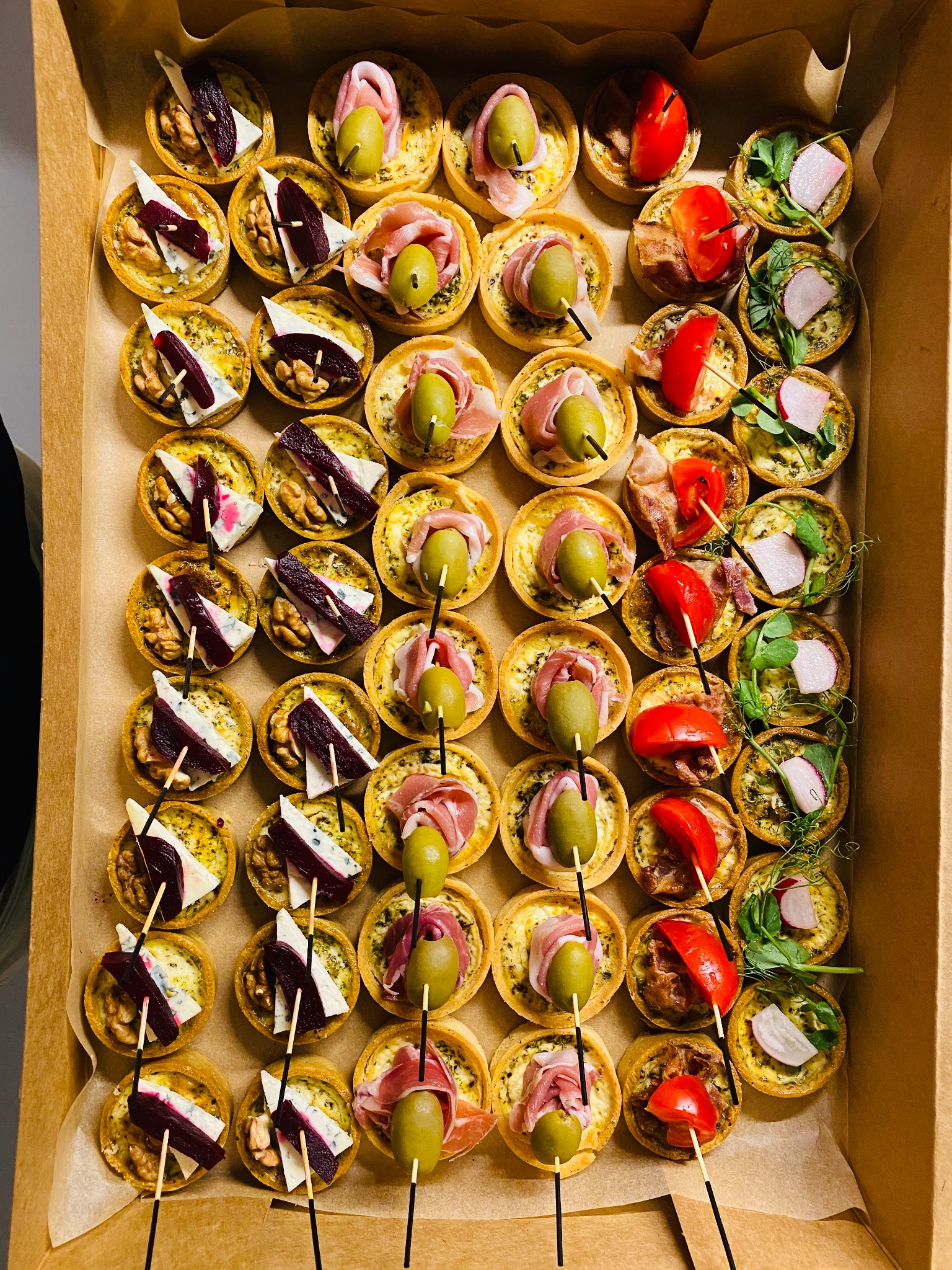 Kartonowe pudełko wypełnione różnorodnymi, małymi tartaletkami na słono. Widoczne tartaletki z burakiem, serem pleśniowym i orzechem włoskim, z szynką parmeńską i oliwką oraz z pomidorem i rzodkiewką.