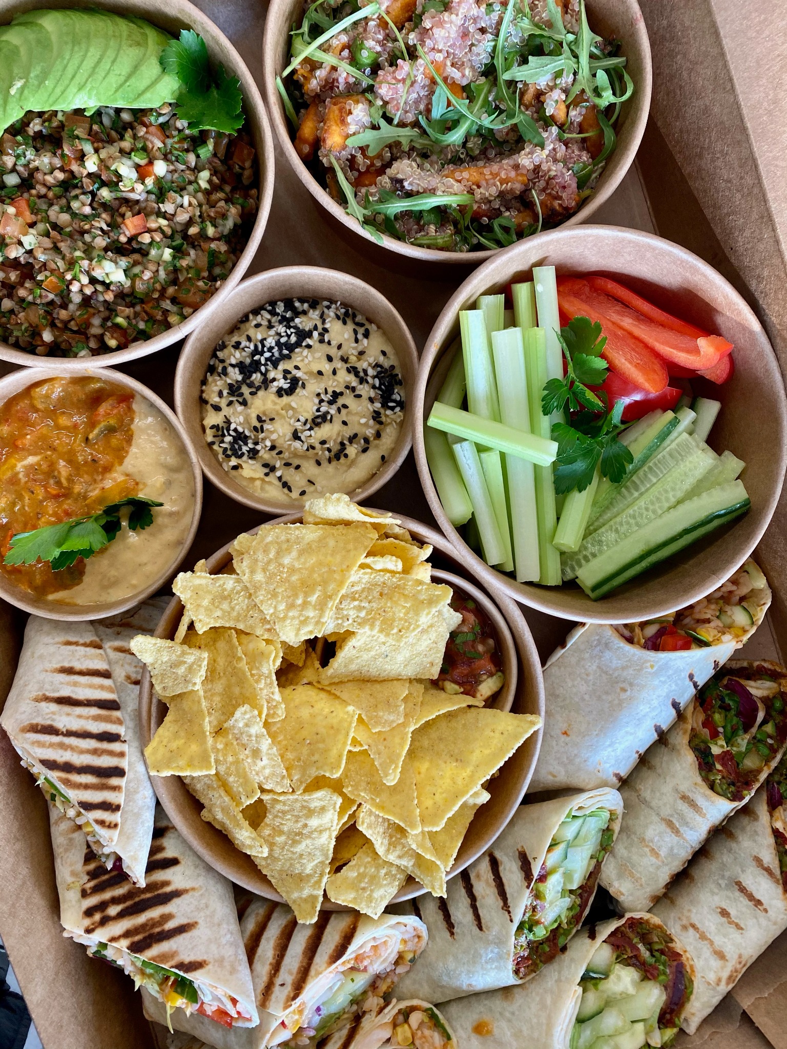 Zestaw różnorodnych dań w papierowych miseczkach i wrapów, w tym sałatka z awokado, quinoa z warzywami, hummus z czarnym sezamem, nachosy z salsą oraz grillowane wrapy z warzywami, ułożonych...