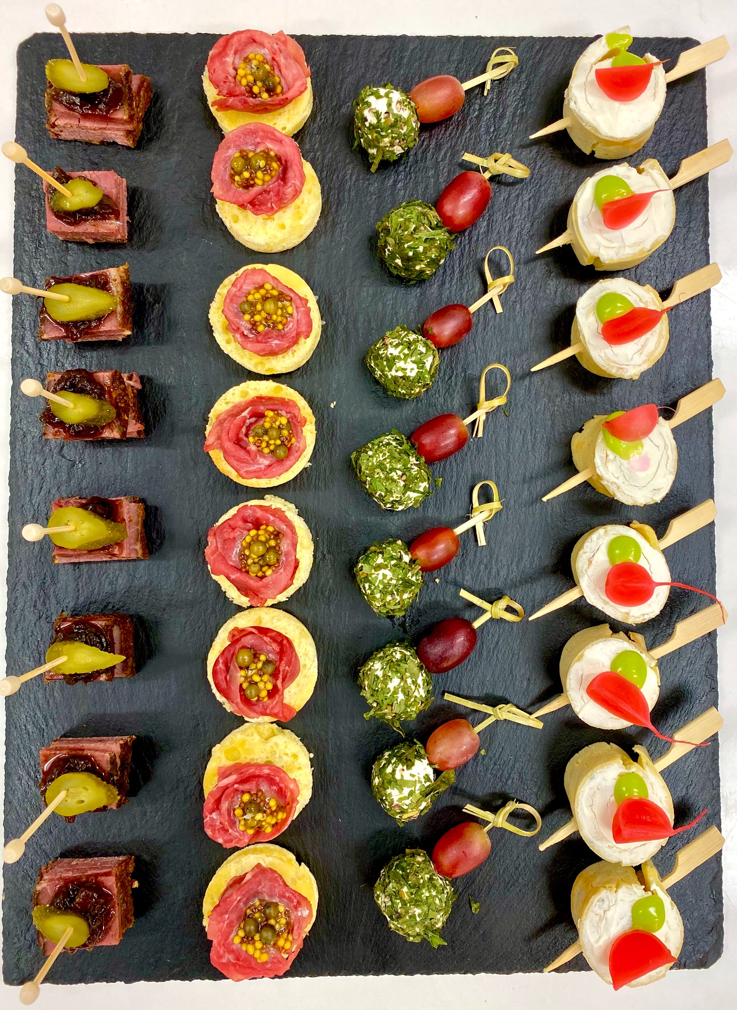 Elegancko ułożone na czarnym kamiennym talerzu różnorodne przekąski cateringowe na drewnianych wykałaczkach: mini kanapeczki z rostbefem i piklami, tartinki z szynką i gorczycą, szaszłyki...