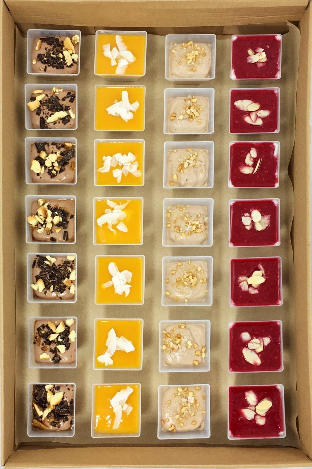 Kartonowe pudełko z 24 małymi, kwadratowymi pojemnikami z deserami o różnych smakach i dekoracjach: czekoladowe z kruszonką, mango z wiórkami kokosowymi, orzechowe z posiekanymi orzechami...