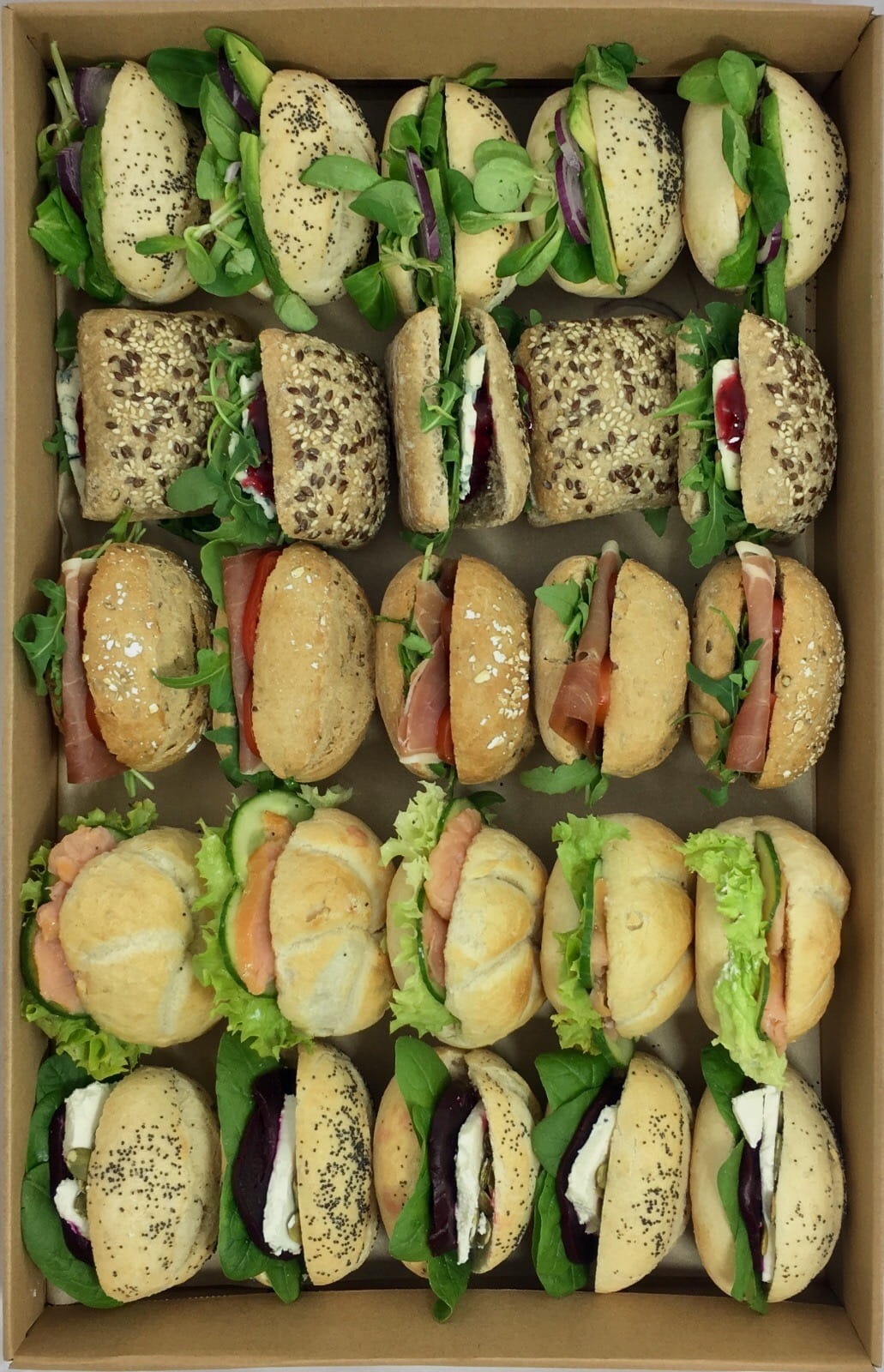 Różnorodne kanapki w kartonowym pudełku, widoczne z góry, prezentujące ofertę cateringową w Poznaniu: kanapki z łososiem, szynką parmeńską, serem pleśniowym i burakiem, avocado i warzywami,...