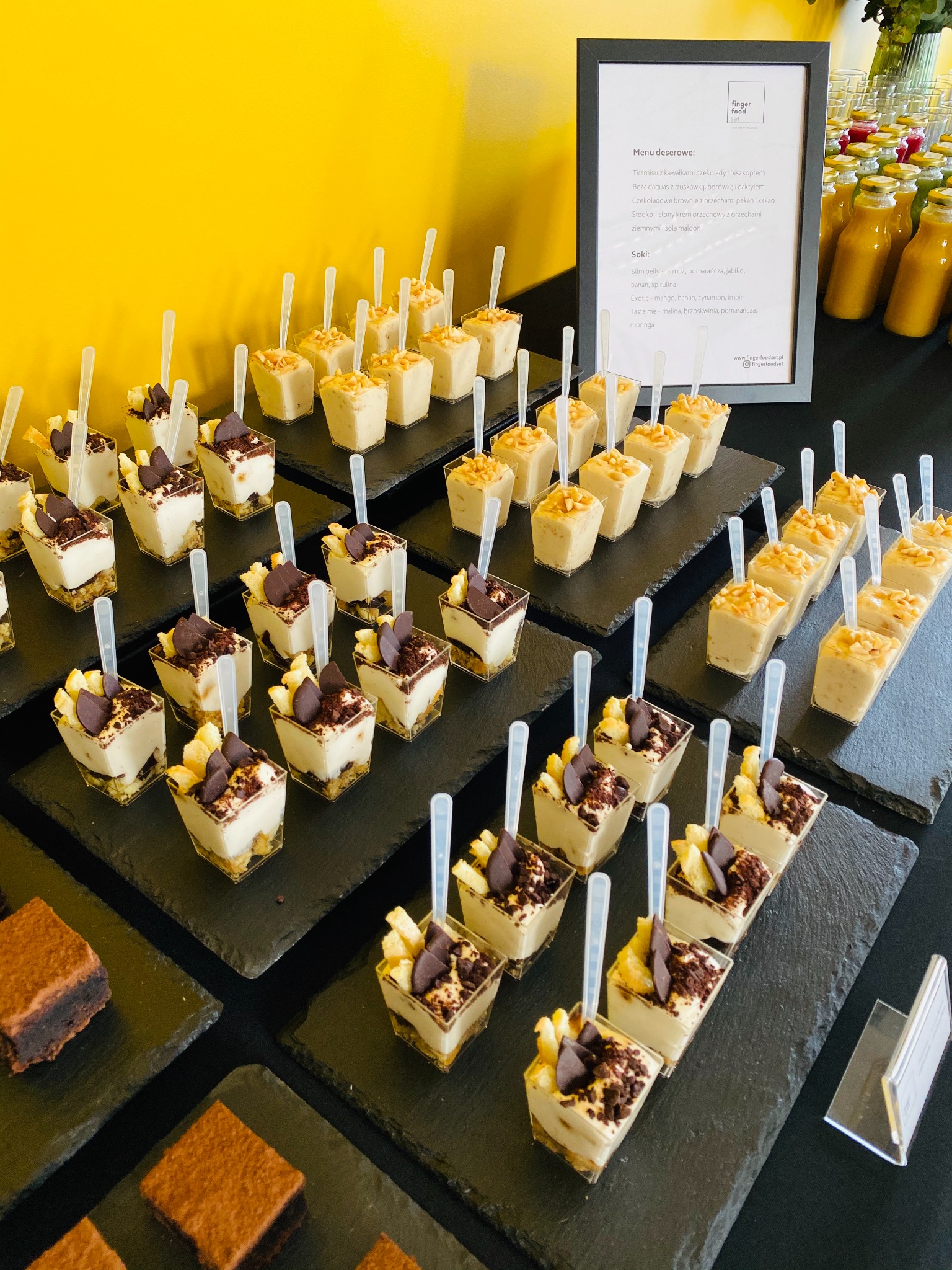 Rzędy deserów w małych plastikowych pojemnikach, ułożone na czarnych łupkowych płytkach, z menu i butelkami soków w tle, prezentacja finger food.