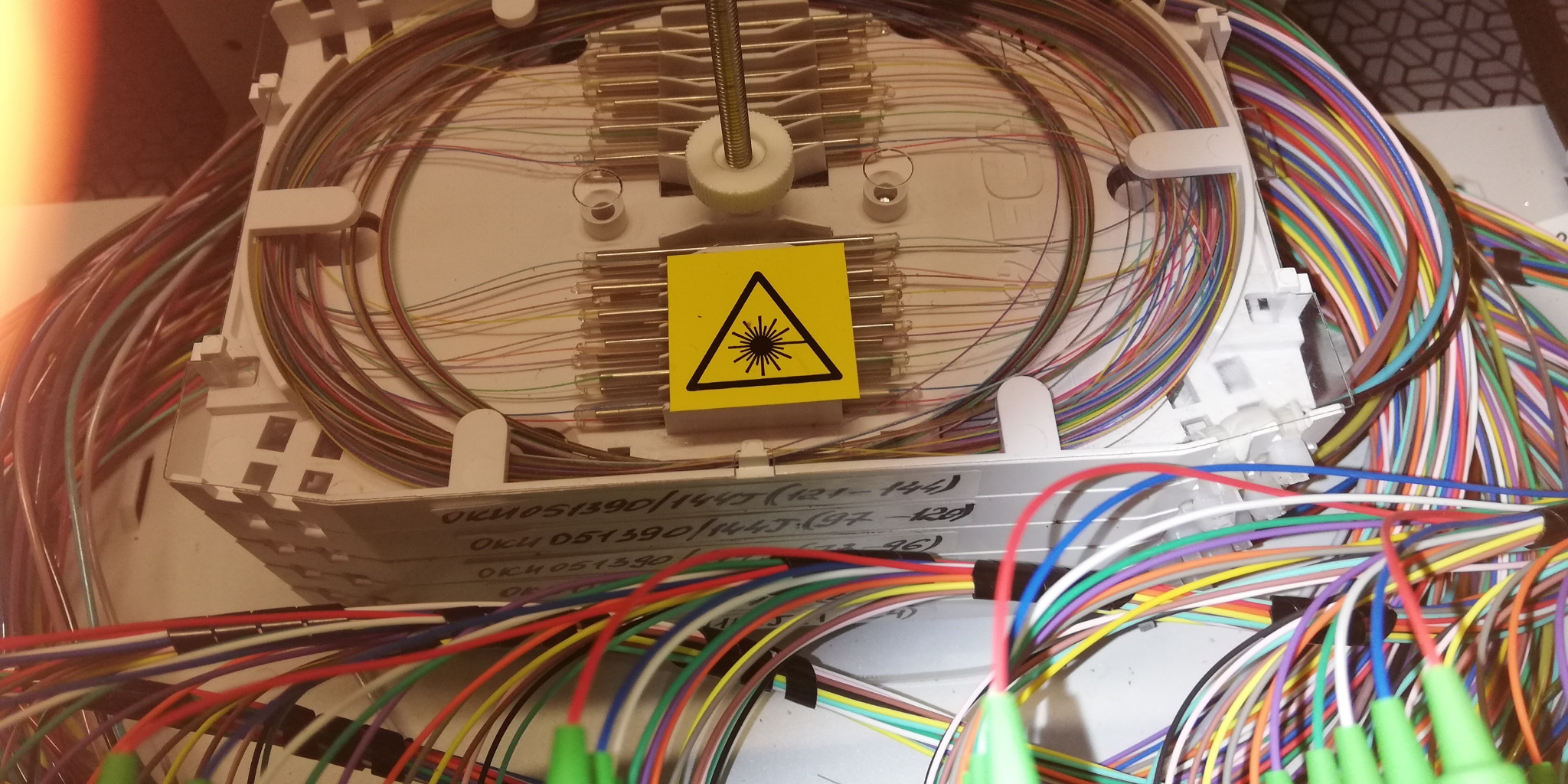 Rozdzielnica światłowodowa z żółtą naklejką ostrzegawczą o promieniowaniu laserowym, widoczne kolorowe kable światłowodowe i oznaczenia na obudowie.