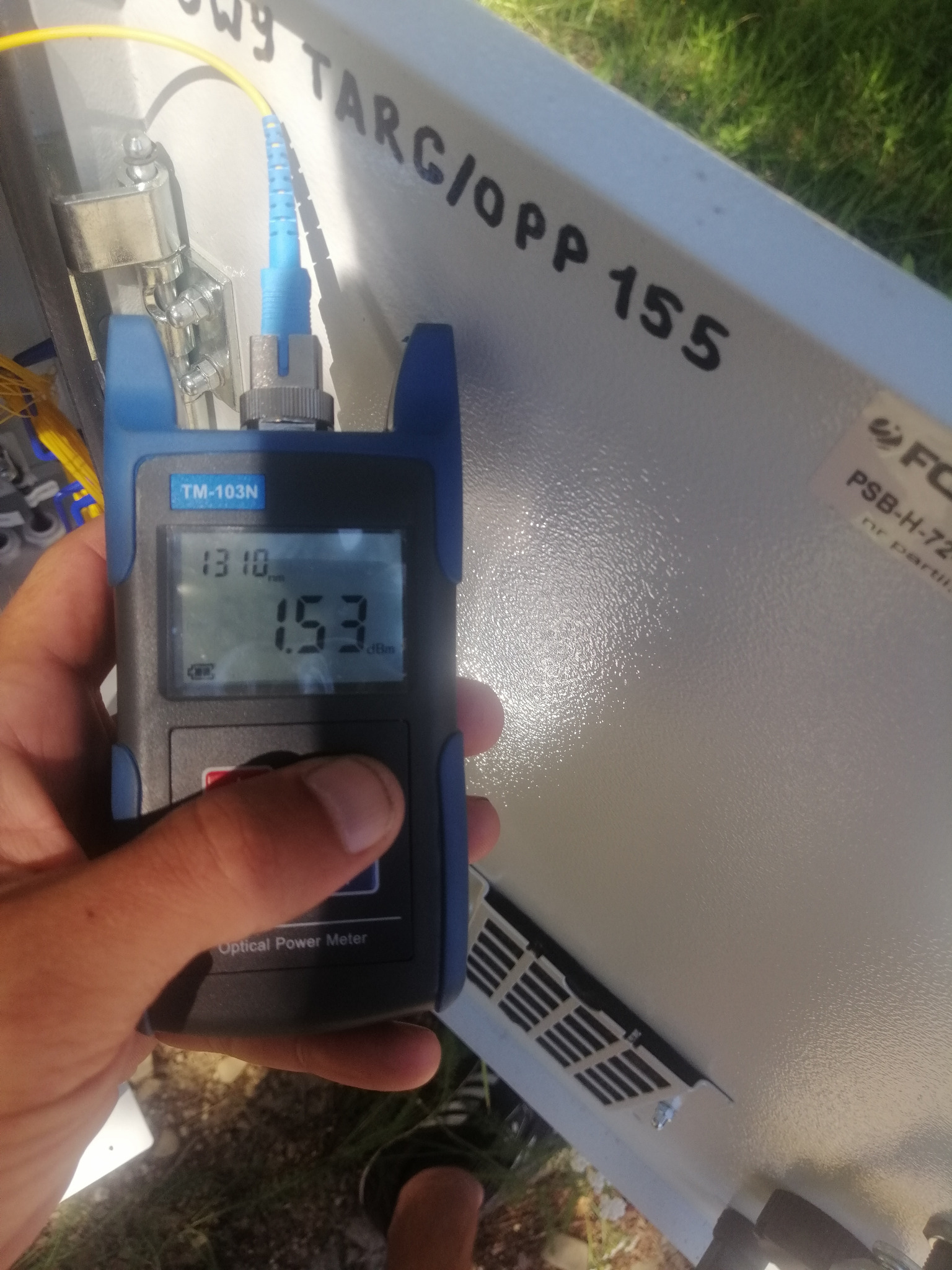 Pomiar mocy sygnału światłowodowego za pomocą miernika TM-103N wewnątrz skrzynki telekomunikacyjnej z napisem 'OWY TARG/OPP 155', widoczny fragment złącza światłowodowego.