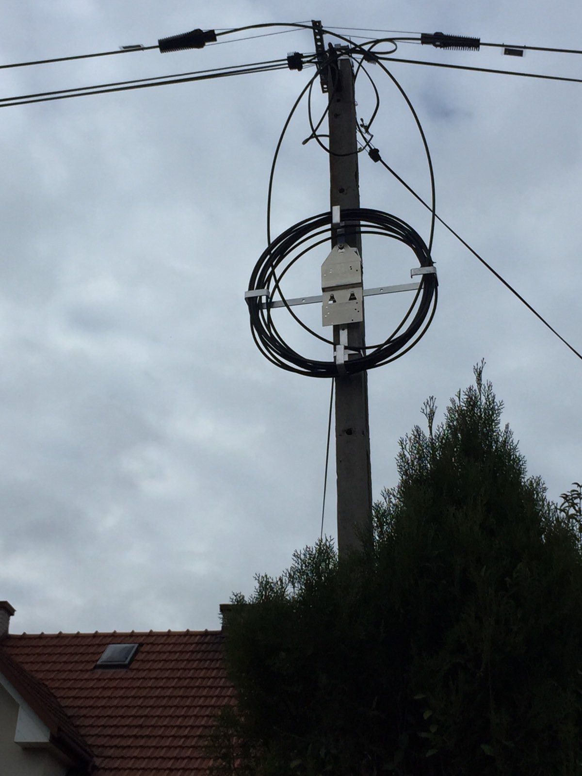 Słup telekomunikacyjny z zamocowanym okablowaniem światłowodowym w pętli, widoczne uchwyty montażowe i przewody podłączeniowe, pochmurne niebo w tle.