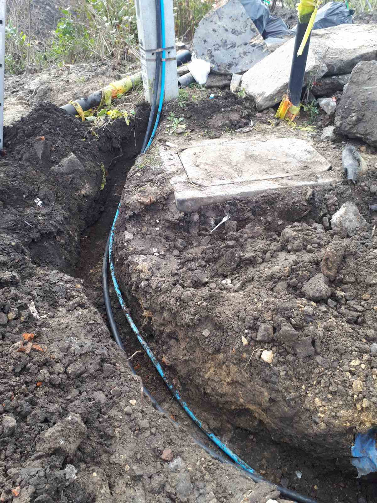 Wykop z ułożonymi kablami telekomunikacyjnymi w niebieskiej i czarnej osłonie, widoczny fragment słupa z przymocowanymi kablami, ziemia z kamieniami, w tle roślinność.