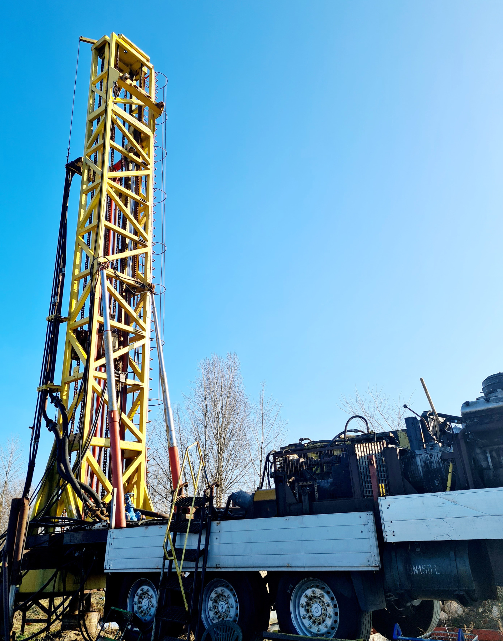 Żółta wieża wiertnicza na platformie ciężarówki z widocznymi elementami silnika i oprzyrządowania na tle błękitnego nieba, sugerująca prace związane z wierceniem studni głębinowych.