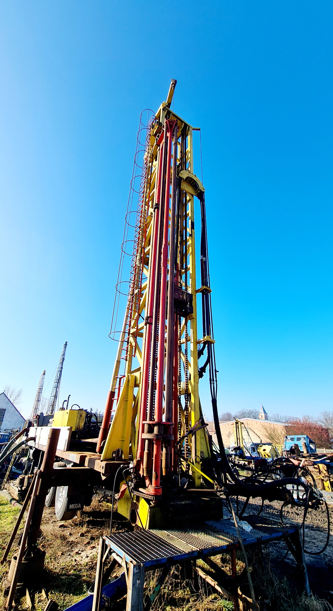 Żółto-czerwona wiertnica ustawiona pionowo na tle błękitnego nieba, widoczna drabina i detale konstrukcyjne, w tle budynki i pojazdy.