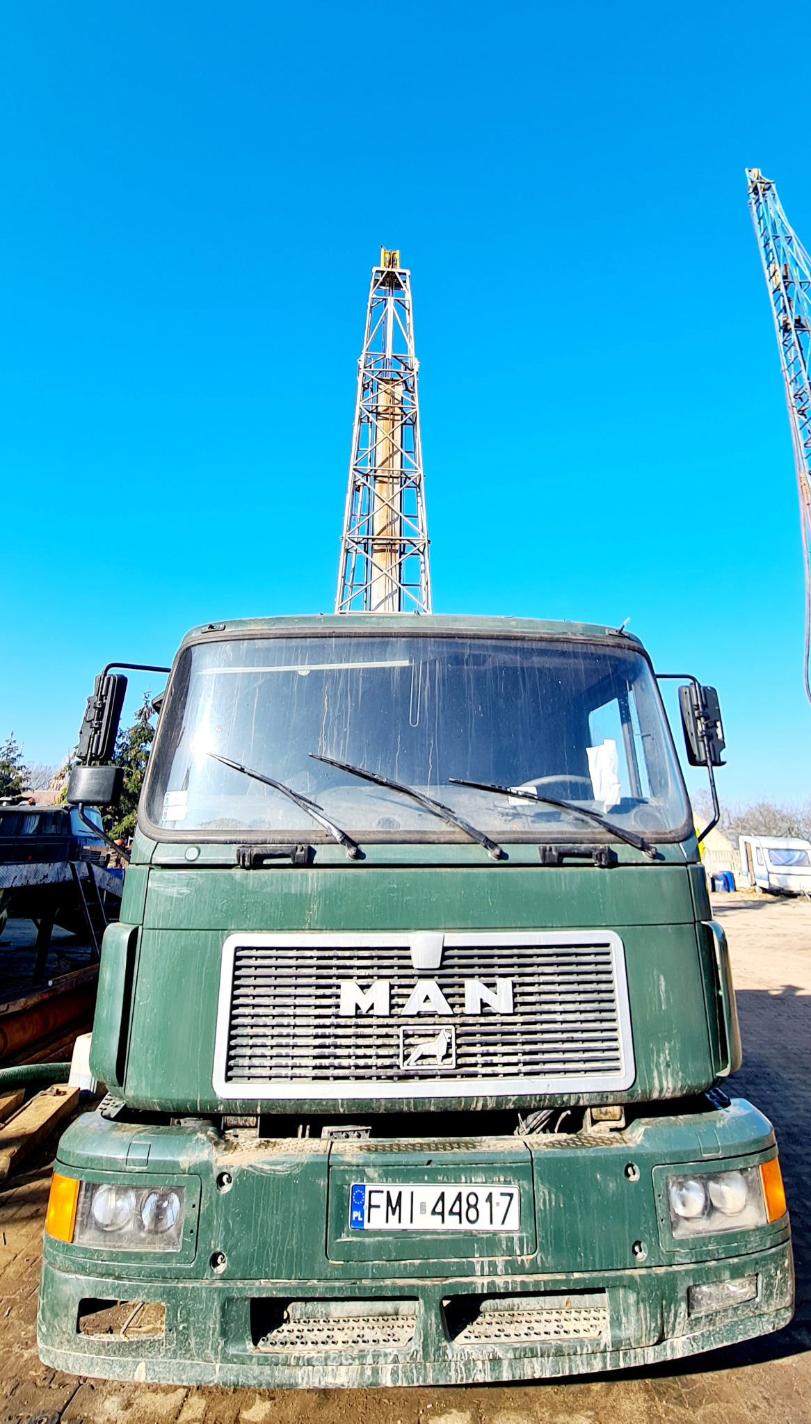 Zielony, brudny samochód ciężarowy MAN na tle wieży wiertniczej i błękitnego nieba, z widoczną tablicą rejestracyjną FMI 44817.