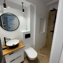 Nowoczesna łazienka z owalną umywalką na drewnianym blacie, czarną armaturą, okrągłym lustrem i kabiną prysznicową wyłożoną płytkami imitującymi drewno, oświetlona czarnymi lampami sufitowymi.