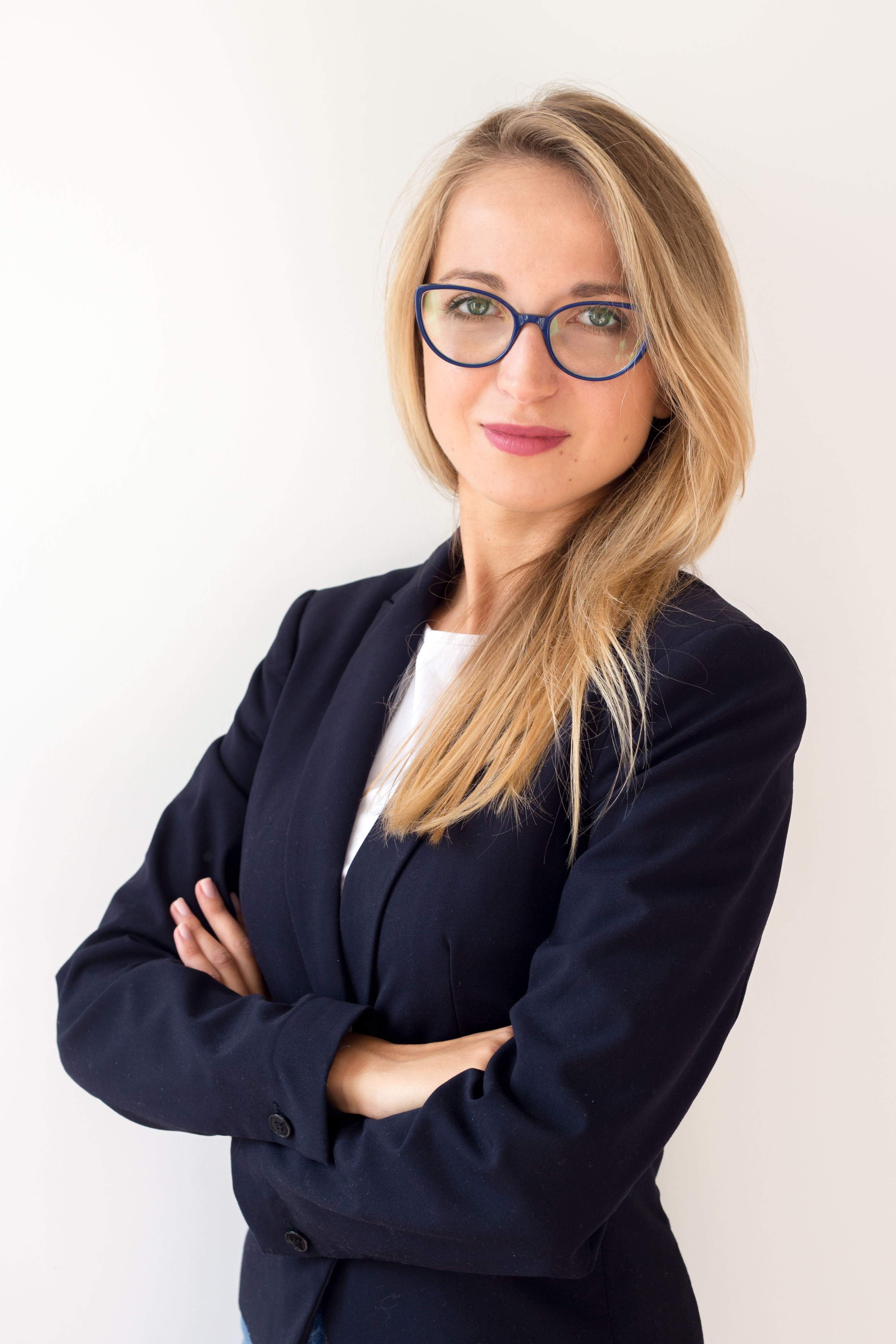 Portret kobiety w granatowej marynarce i okularach o niebieskich oprawkach, z blond włosami opadającymi na ramiona, stojącej na tle białej ściany.