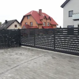 Nowoczesna, przesuwna brama wjazdowa z poziomymi panelami w kolorze grafitowym, uzupełniona furtką, na tle kostki brukowej i budynków mieszkalnych.
