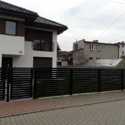 Nowoczesne, czarne ogrodzenie metalowe poziome z furtką, na tle jasnego domu z elementami drewnopodobnymi i kostki brukowej.