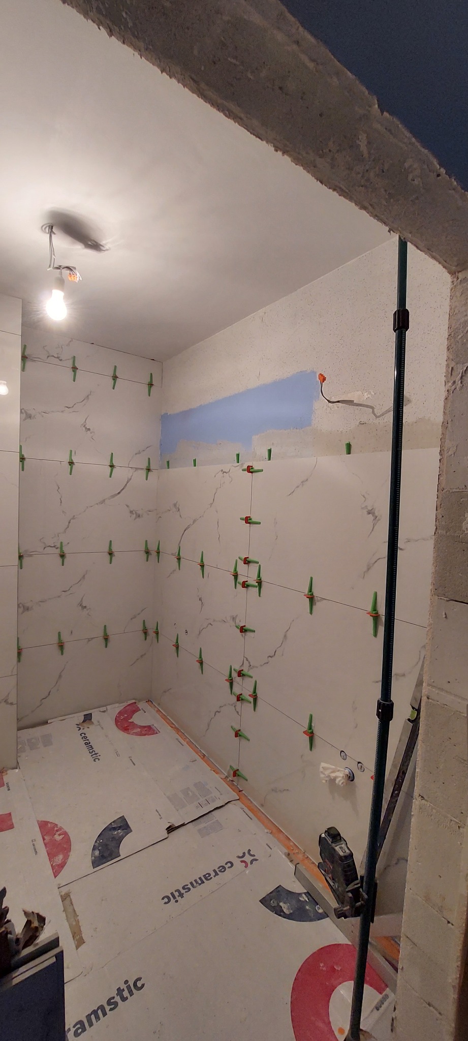 Łazienka w trakcie układania płytek imitujących marmur, z użyciem klipsów poziomujących, widoczne narzędzia glazurnicze i częściowo pomalowana ściana na niebiesko.