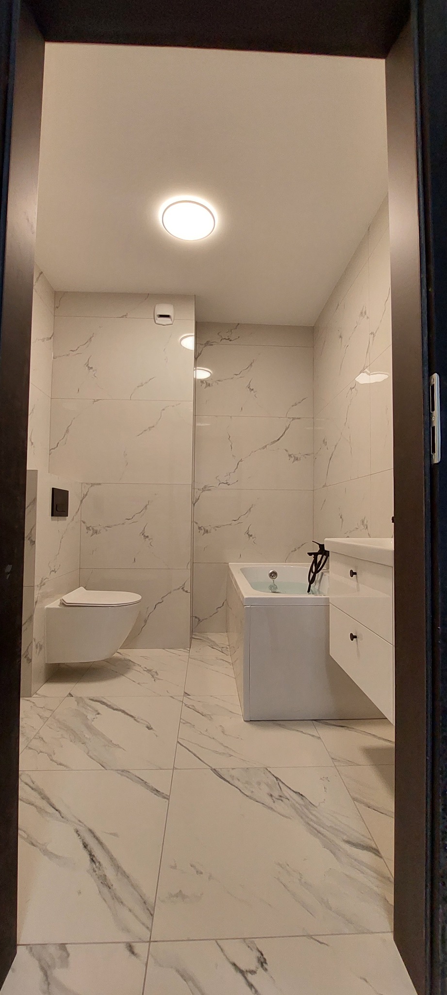 Nowoczesna łazienka z białymi płytkami imitującymi marmur, wiszącą toaletą, prostokątną wanną z czarną armaturą i szafką z umywalką, oświetlona centralnym plafonem.