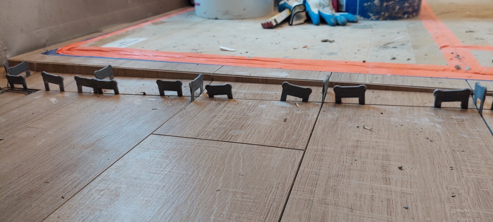 Układanie płytek podłogowych z użyciem systemu poziomowania; widoczne klipsy i kliny, w tle wiadro, rękawice robocze i zabezpieczenie podłogi.