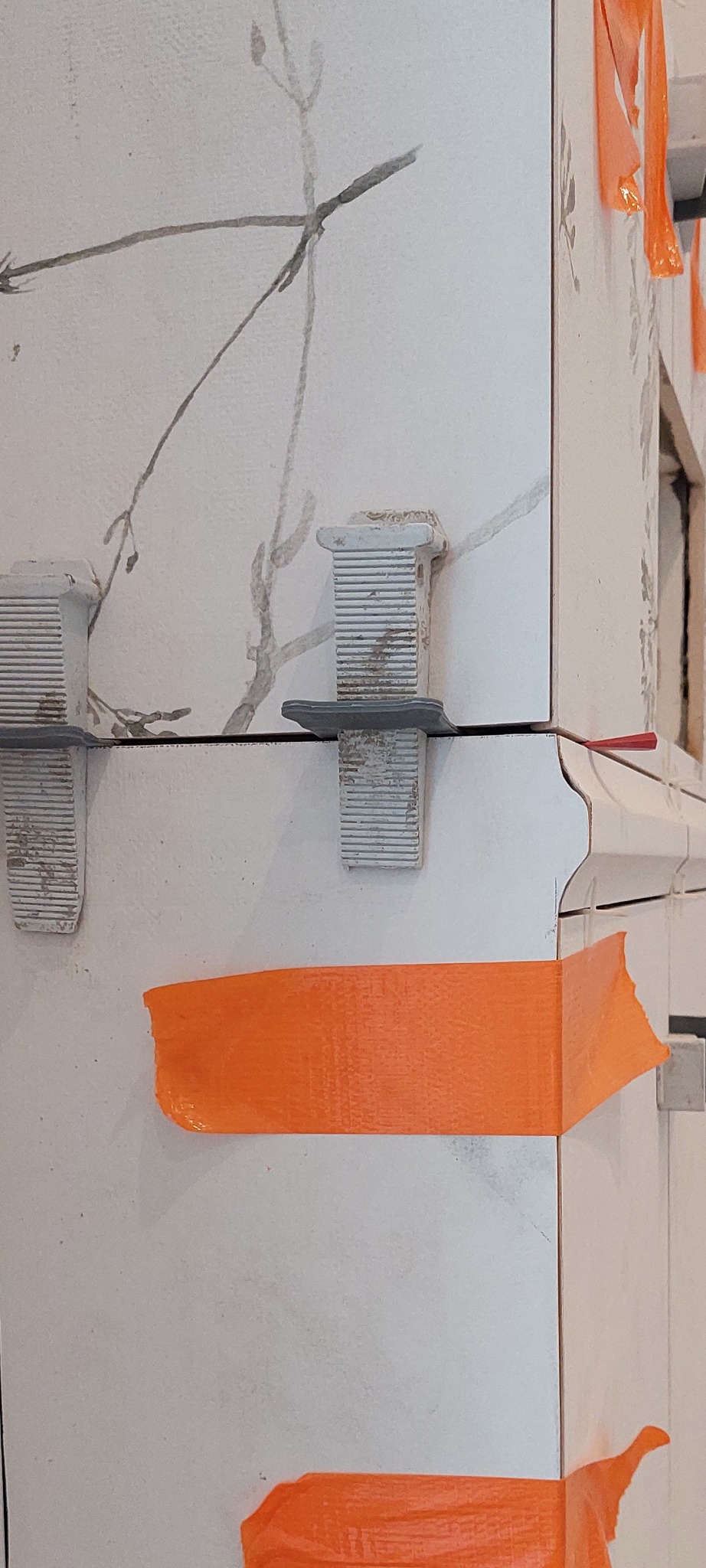 Fragment ściany z białymi płytkami ceramicznymi, wzór imitujący gałązki, zabezpieczonymi pomarańczową taśmą malarską i poziomującymi klipsami do płytek.